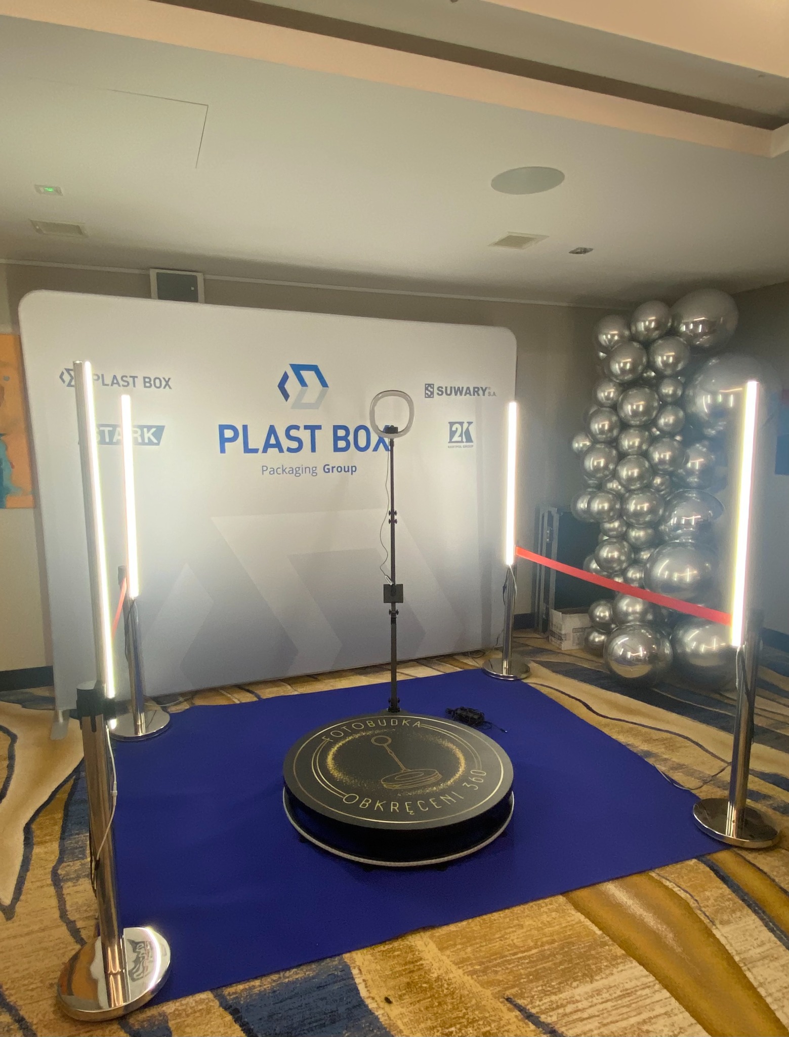 Rozstawiona platforma obrotowa 360 stopni z napisem 'Fotobudka' i oświetleniem LED, ustawiona na niebieskim dywanie, z tłem reklamowym firm i dekoracją z srebrnych balonów.