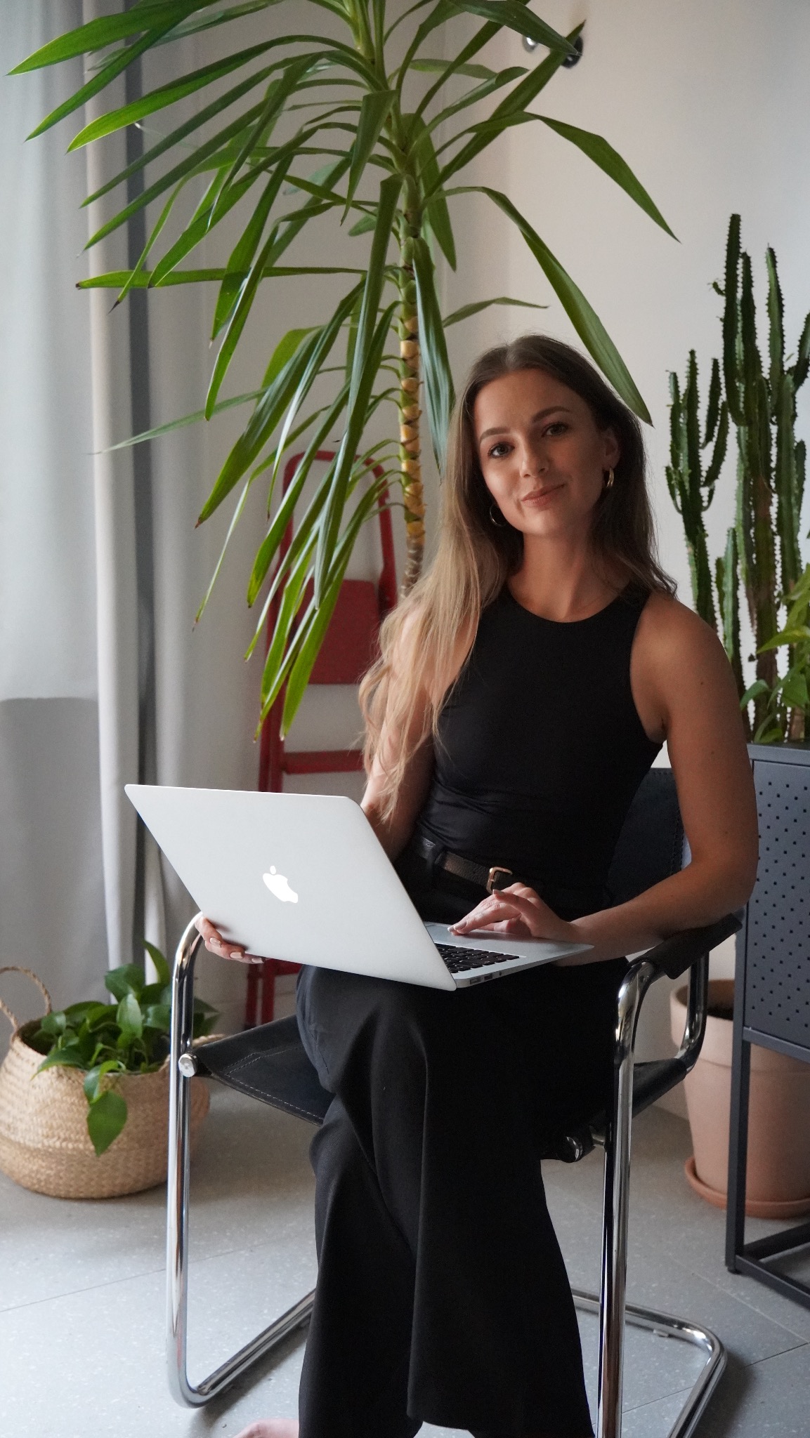 Kobieta w czarnym stroju siedzi na krześle z laptopem, otoczona roślinami doniczkowymi, w tle czerwona drabina.