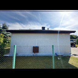 Handy Man - Biały budynek gospodarczy z brązowym dachem, świeżo po malowaniu, zabezpieczony taśmą malarską, widoczny przez zielone ogrodzenie z siatki.