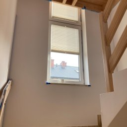 Handy Man - Świeżo pomalowana ściana klatki schodowej na biało, zabezpieczona taśmą malarską przy oknie i drewnianej konstrukcji sufitu, widoczne płytki imitujące drewno na schodach.