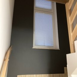 Handy Man - Ściana pomalowana na ciemny kolor obok schodów wyłożonych płytkami imitującymi drewno, z oknem z żaluzjami