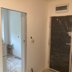 Handy Man - Wnętrze w trakcie odświeżania: świeżo pomalowane ściany na biało, zabezpieczone folią drzwi, wiadro z farbą i pędzlem przy ścianie, widoczne ślady prac na podłodze.