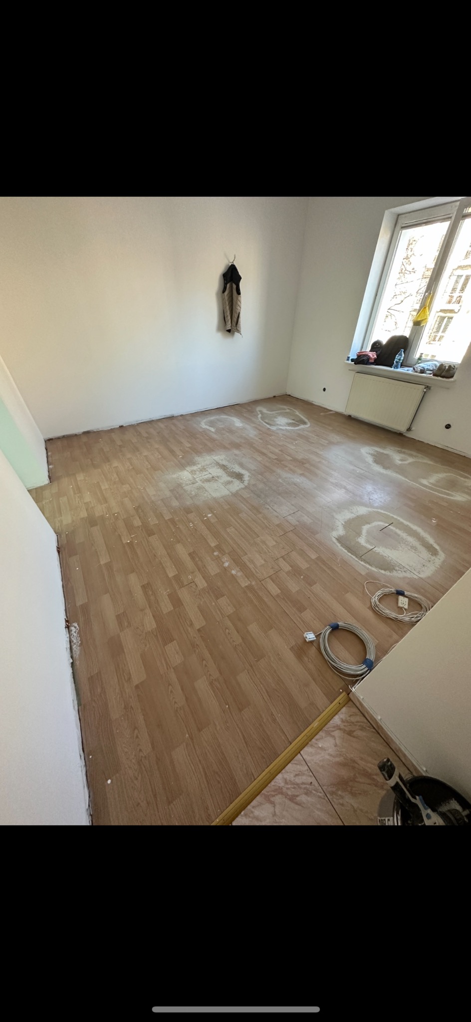 Pokój z częściowo ułożoną podłogą z paneli imitujących drewno, widoczne ślady po wcześniejszej pracy, białe ślady na panelach, w rogu zwinięte kable elektryczne, listwa przypodłogowa jeszcze nie...