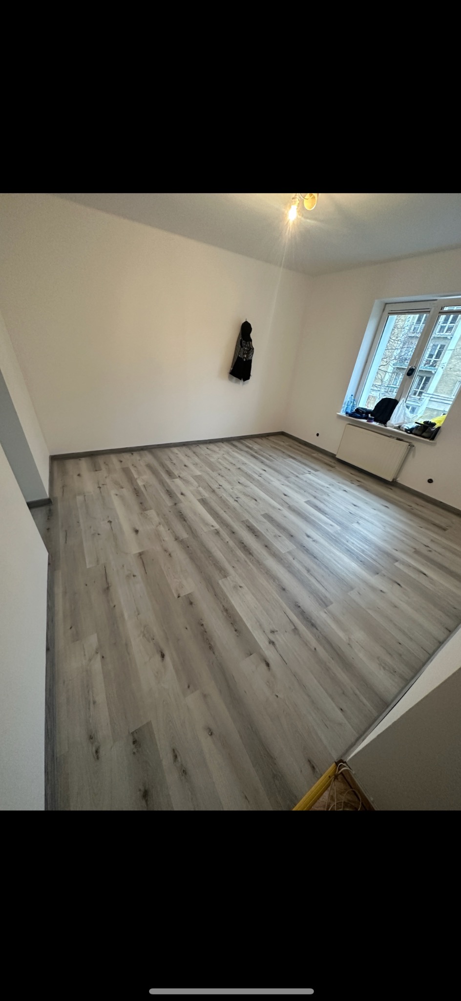 Pusty pokój po remoncie z nową, jasną podłogą z paneli imitujących drewno; kurtka zawieszona na ścianie, grzejnik pod oknem.