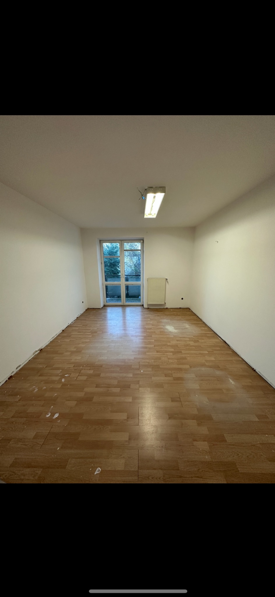 Pusty pokój po remoncie z panelami podłogowymi, ślady farby na podłodze, świeżo pomalowane ściany na biało, okno balkonowe z widokiem na drzewa.