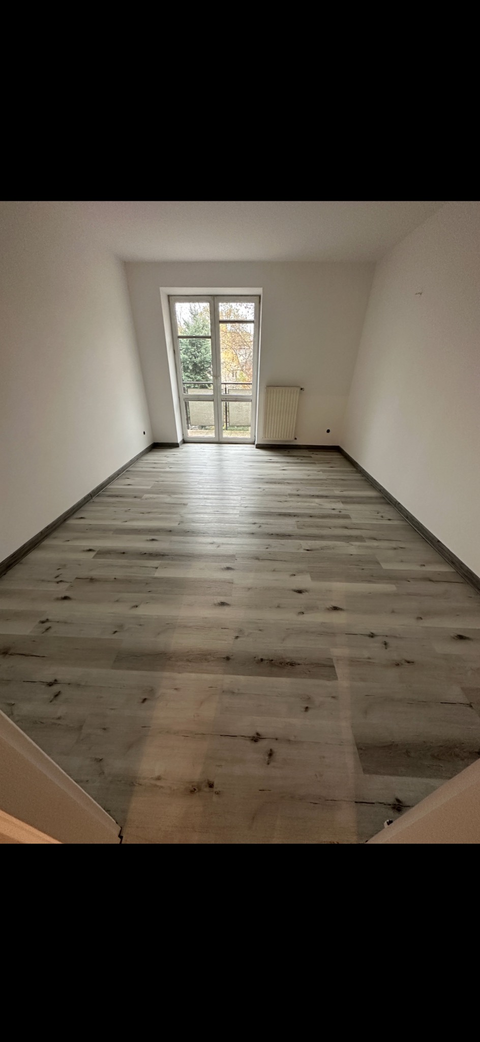 Pusty pokój na poddaszu z nową podłogą z paneli winylowych imitujących drewno, widok na balkon i grzejnik