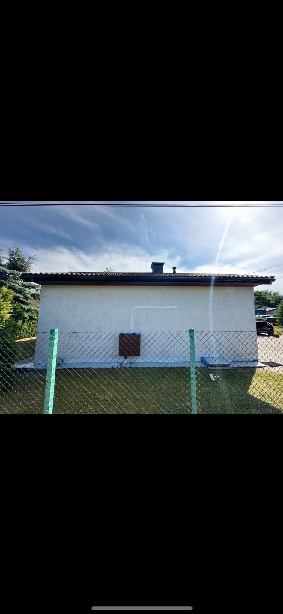 Biały budynek gospodarczy z brązowym dachem, świeżo po malowaniu, zabezpieczony taśmą malarską, widoczny przez zielone ogrodzenie z siatki.