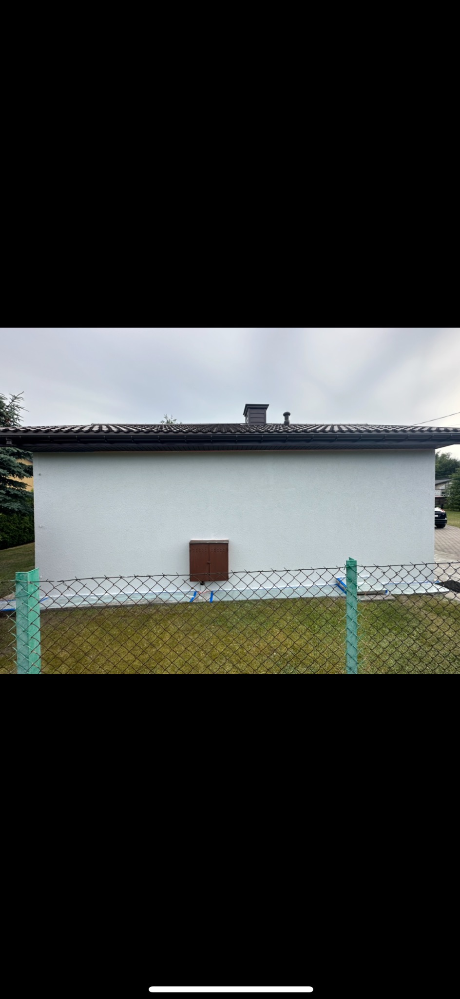 Świeżo pomalowana elewacja budynku jednokondygnacyjnego z brązową skrzynką pocztową, zabezpieczona folią malarską przy gruncie, widoczna zza ogrodzenia z siatki.