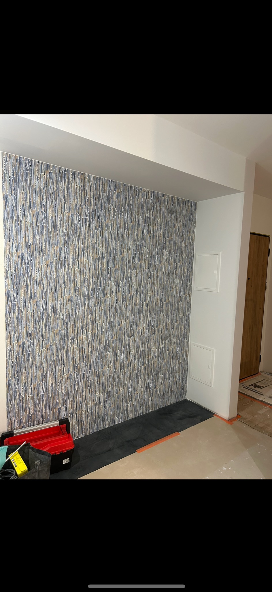 Ściana wykończona tapetą z motywem liści w odcieniach szarości i beżu, widoczna skrzynka z narzędziami i fragment podłogi zabezpieczony czarną folią.