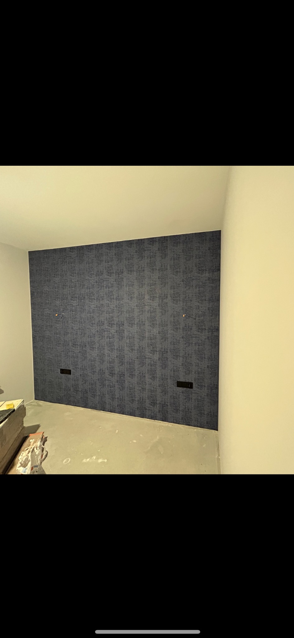 Ściana w pokoju po tapetowaniu, widoczne gniazdka elektryczne, surowy beton na podłodze i materiały budowlane w rogu pomieszczenia.