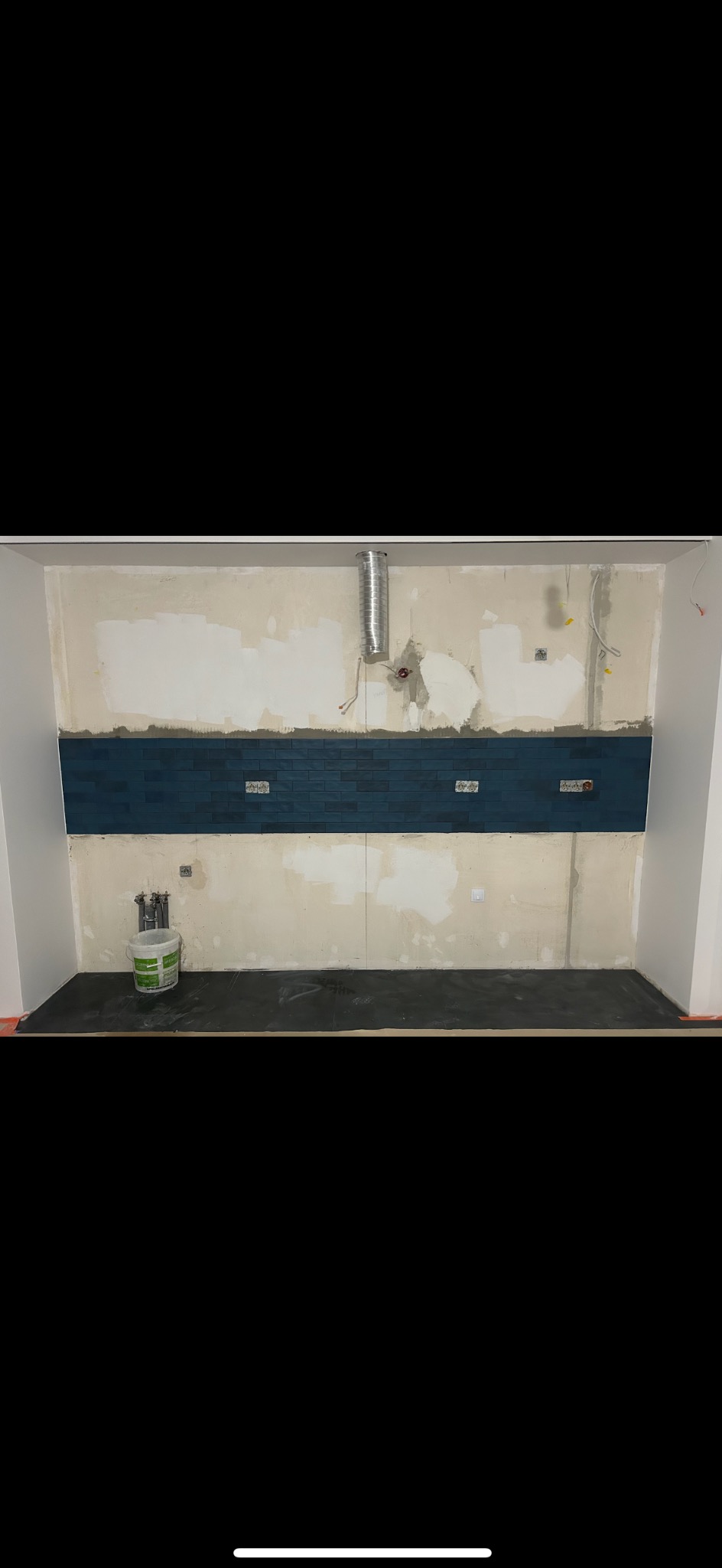 Fragment ściany z częściowo ułożonymi, ciemnoniebieskimi płytkami w kształcie cegiełek, widoczne puszki elektryczne, ślady po kleju i instalacja wentylacyjna, podłoga zabezpieczona folią, wiadro...