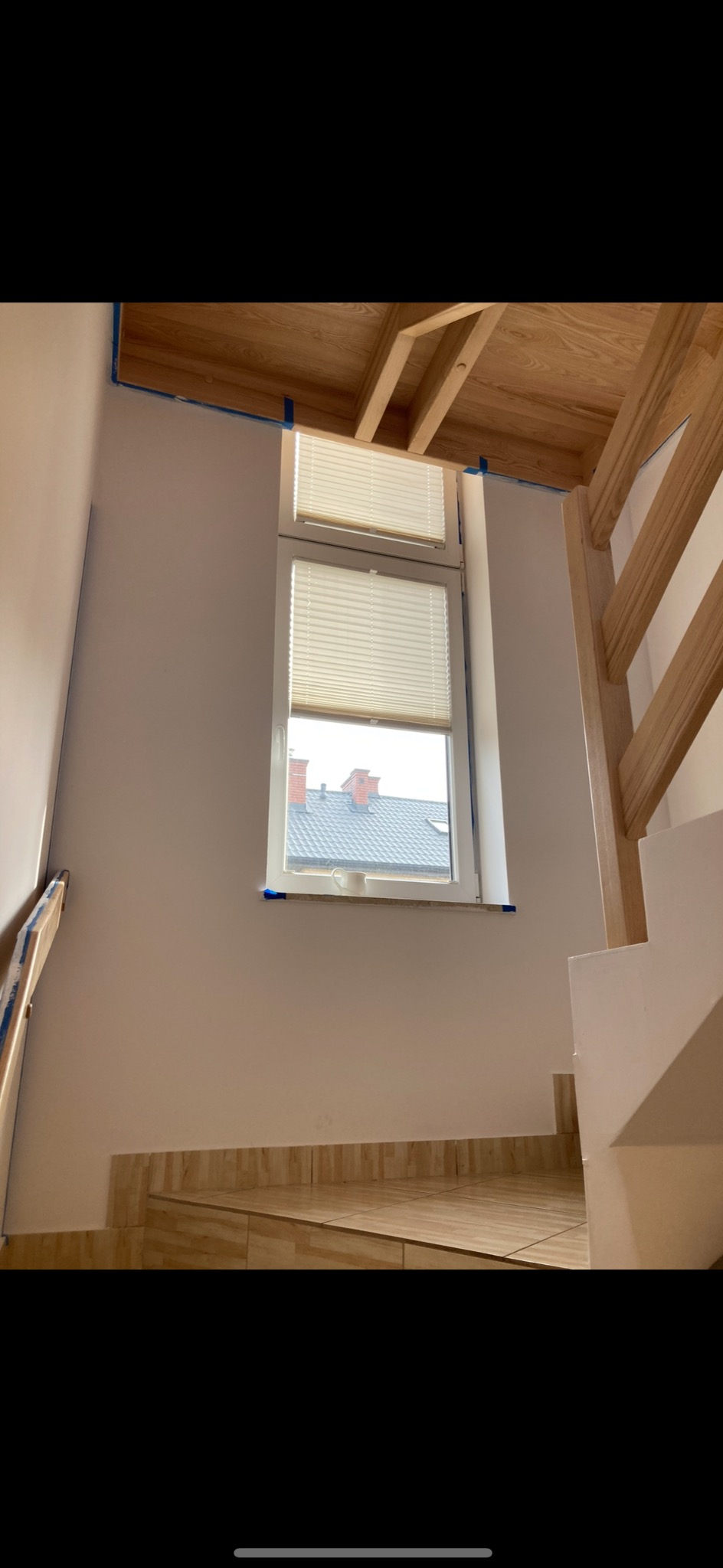 Świeżo pomalowana ściana klatki schodowej na biało, zabezpieczona taśmą malarską przy oknie i drewnianej konstrukcji sufitu, widoczne płytki imitujące drewno na schodach.