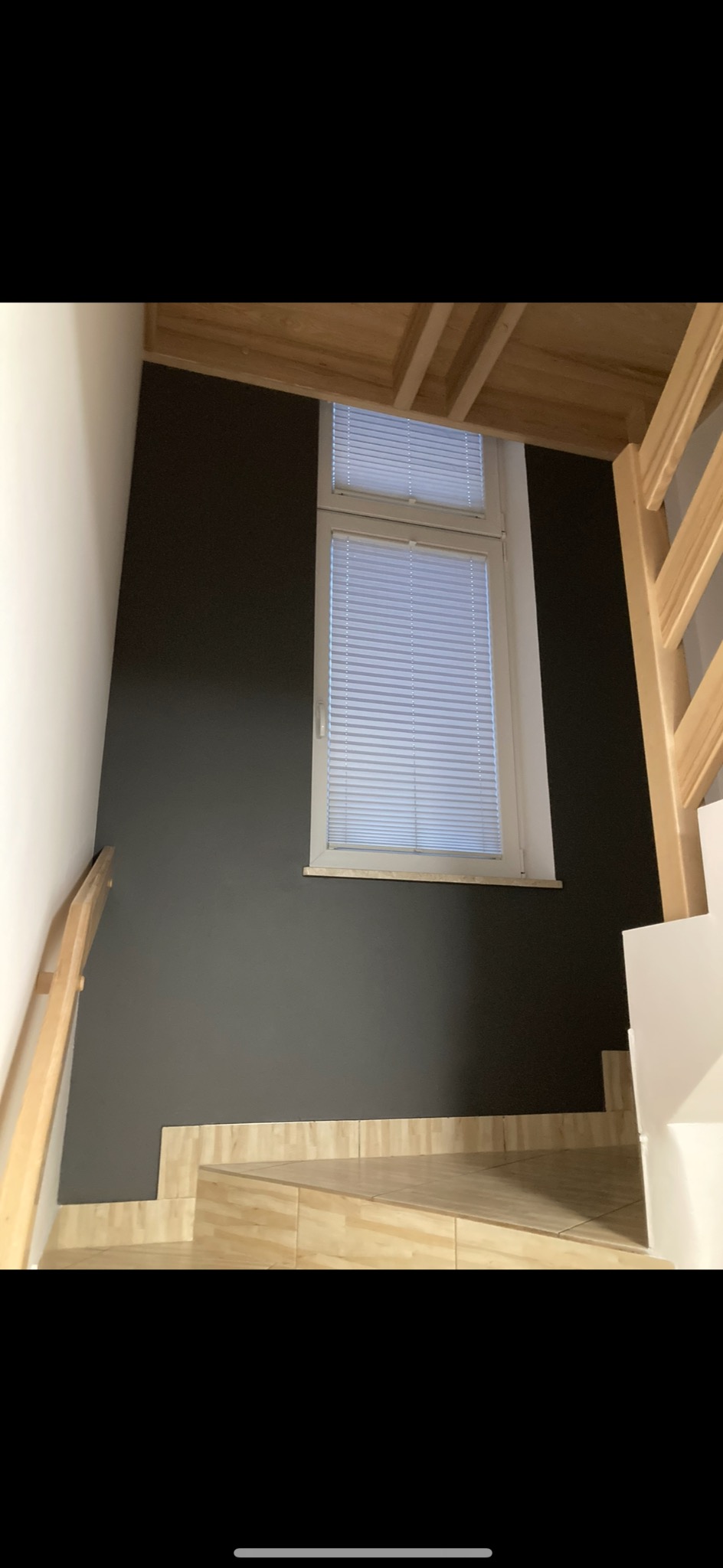 Ściana pomalowana na ciemny kolor obok schodów wyłożonych płytkami imitującymi drewno, z oknem z żaluzjami