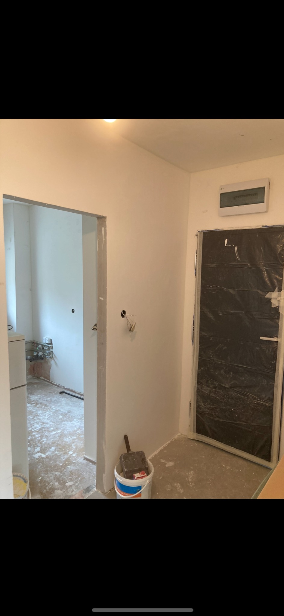 Wnętrze w trakcie odświeżania: świeżo pomalowane ściany na biało, zabezpieczone folią drzwi, wiadro z farbą i pędzlem przy ścianie, widoczne ślady prac na podłodze.