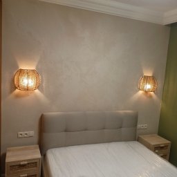 DAW-BUD Dawid Szypulski - Sypialnia po odświeżeniu: beżowe ściany z dekoracyjną fakturą, łóżko z jasnym zagłówkiem zabezpieczone folią, szafki nocne z rattanowymi detalami i plecione lampy ścienne po obu stronach łóżka.