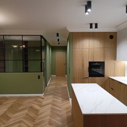 DAW-BUD Dawid Szypulski - Nowoczesne wnętrze z parkietem ułożonym w jodełkę, zielonymi ścianami, zabudową kuchenną z drewnianymi frontami i wyspą z blatem imitującym marmur, oddzielone szklaną ścianą z czarnymi szprosami.