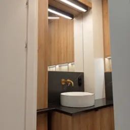 Nowoczesna łazienka z umywalką nablatową, złotą armaturą i oświetleniem LED, widziana zza uchylonych drzwi.