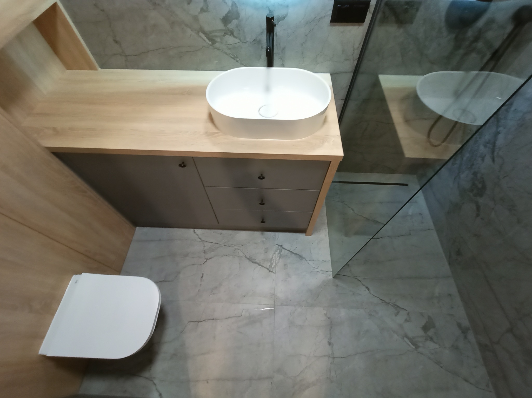 Nowoczesna łazienka z marmurową posadzką, wisząca toaleta, umywalka nablatowa na szafce z szufladami i kabina prysznicowa ze szklaną ścianką.