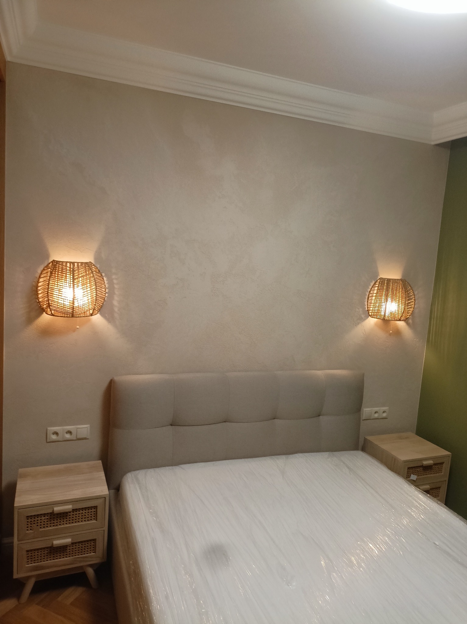 Sypialnia po odświeżeniu: beżowe ściany z dekoracyjną fakturą, łóżko z jasnym zagłówkiem zabezpieczone folią, szafki nocne z rattanowymi detalami i plecione lampy ścienne po obu stronach łóżka.