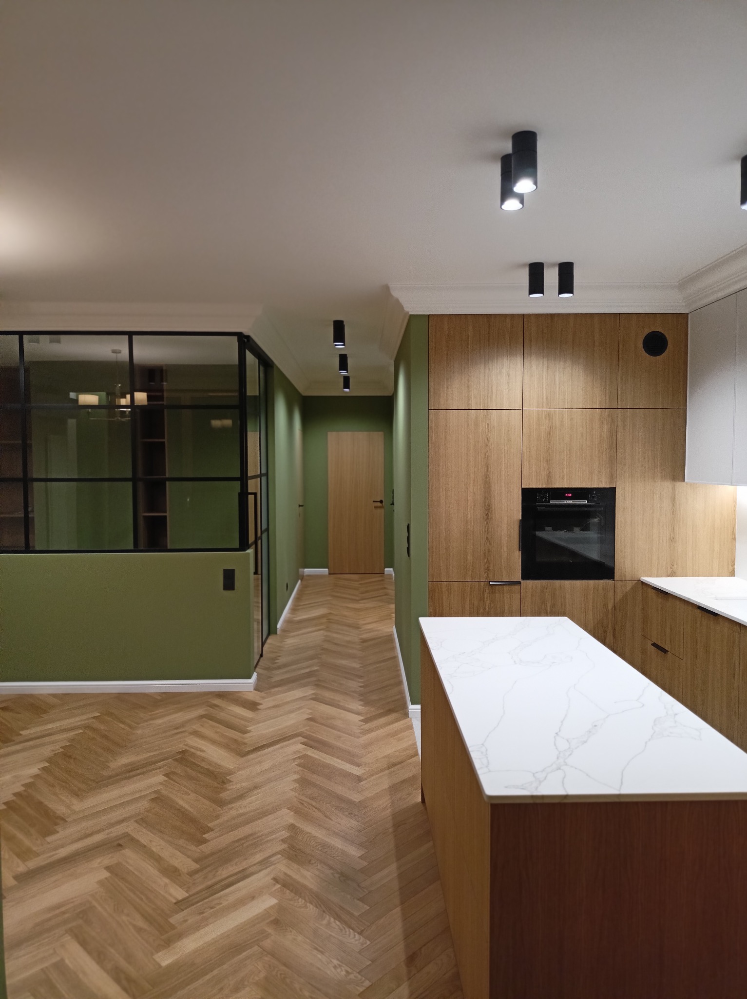 Nowoczesne wnętrze z parkietem ułożonym w jodełkę, zielonymi ścianami, zabudową kuchenną z drewnianymi frontami i wyspą z blatem imitującym marmur, oddzielone szklaną ścianą z czarnymi szprosami.