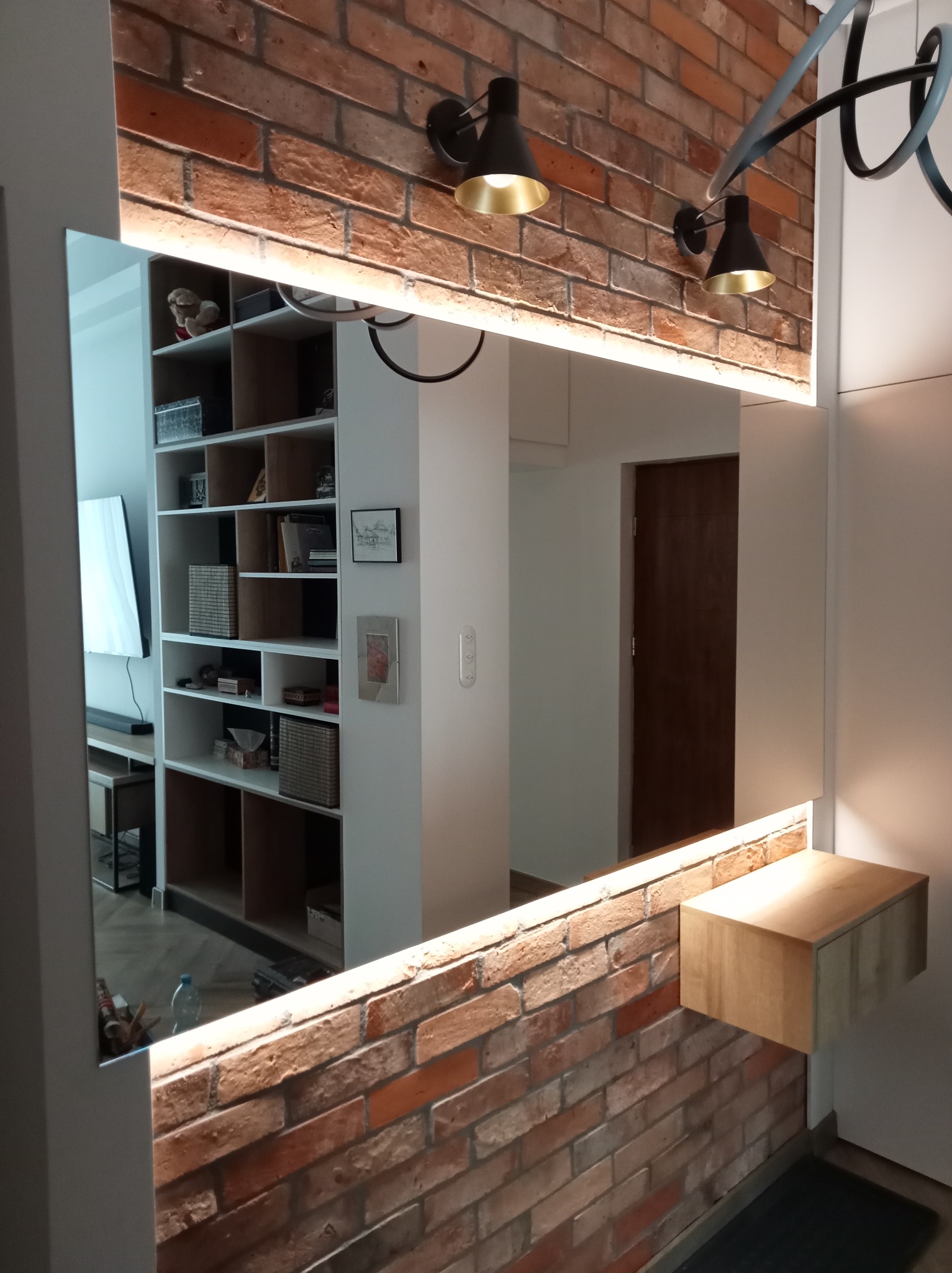 Lustro z oświetleniem LED wkomponowane w ścianę z cegły, z drewnianą półką boczną, odbijające wnętrze mieszkania z regałem i drzwiami.