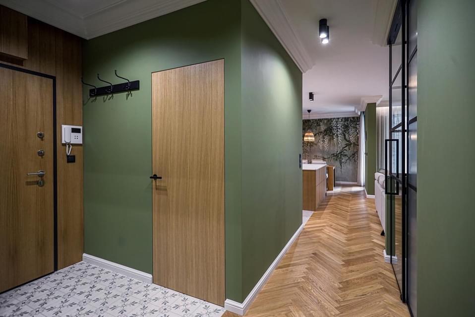 Nowoczesny przedpokój z drewnianą podłogą ułożoną w jodełkę, zielone ściany i ukryte drzwi z drewnianą okleiną, widok na salon z dekoracyjną tapetą.