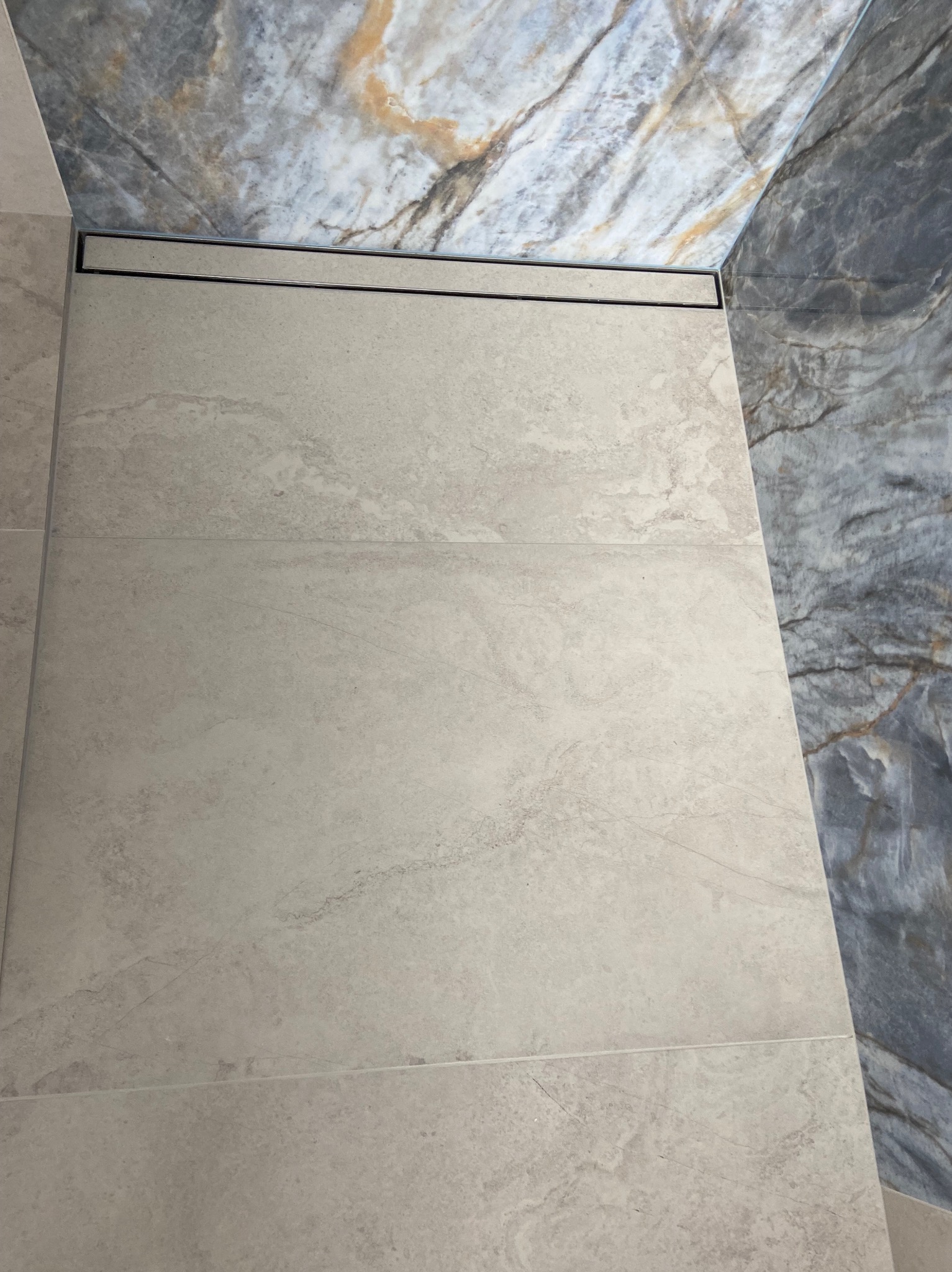 Beżowe płytki podłogowe w łazience, z widocznym odpływem liniowym oraz marmurową okładziną ścienną w odcieniach szarości i beżu.