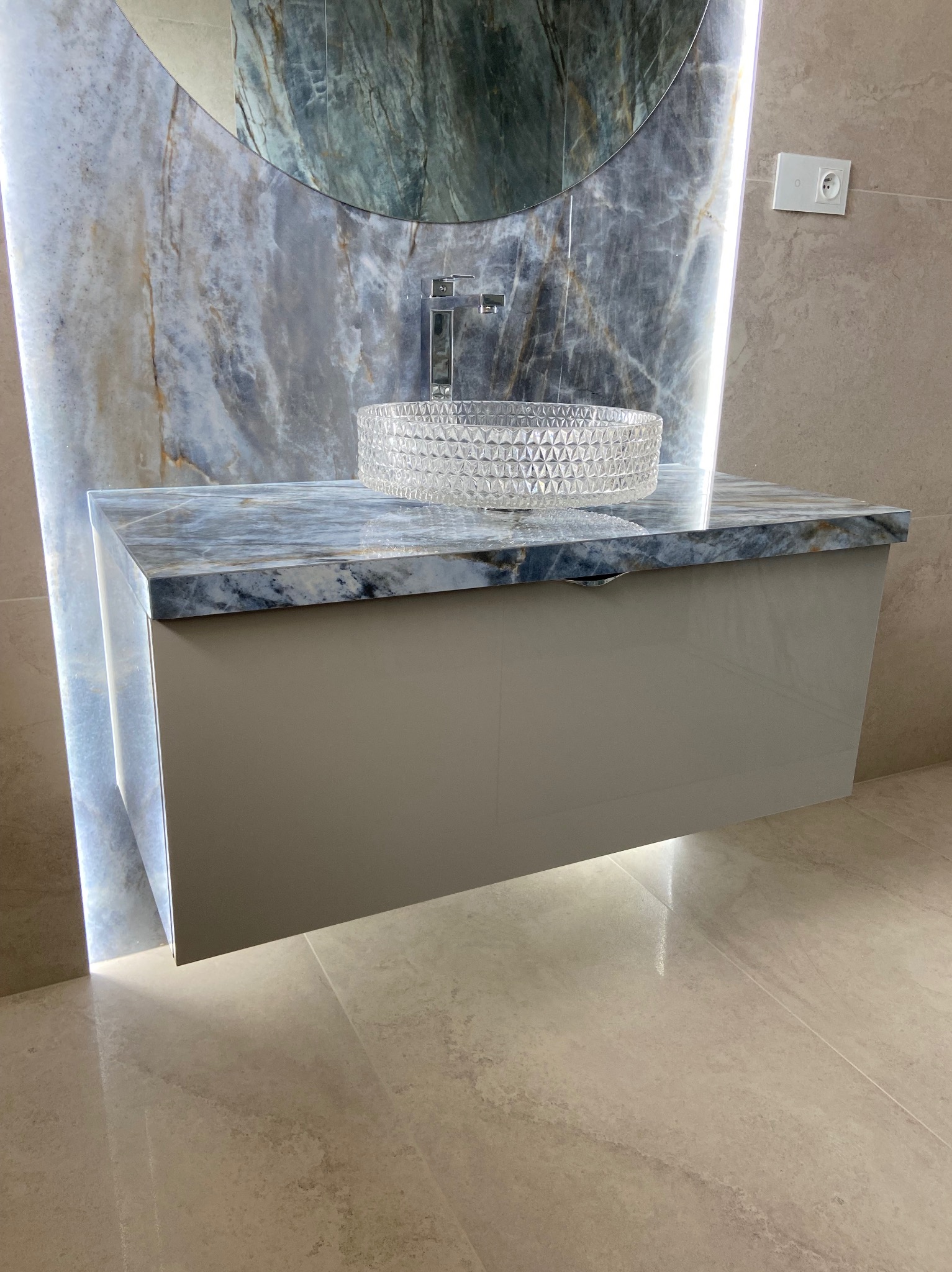 Nowoczesna łazienka z umywalką kryształową na blacie z marmuru, podświetlona listwą LED za lustrem, beżowe płytki podłogowe i ścienne.