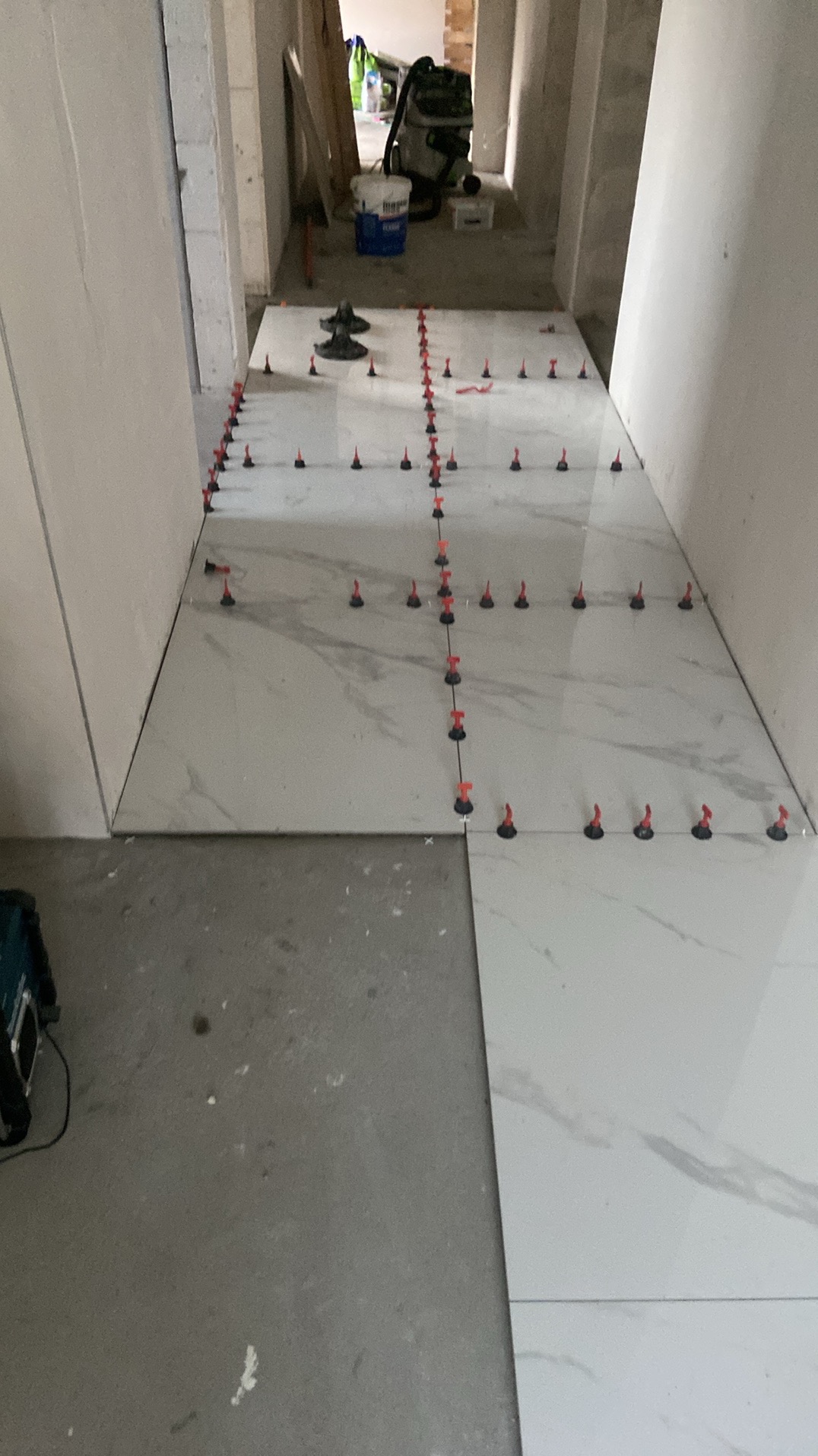 Układanie dużych, białych płytek podłogowych z szarym marmurkowym wzorem, z użyciem systemu poziomowania z czerwonymi klinami, widoczne narzędzia i odkurzacz przemysłowy w tle.