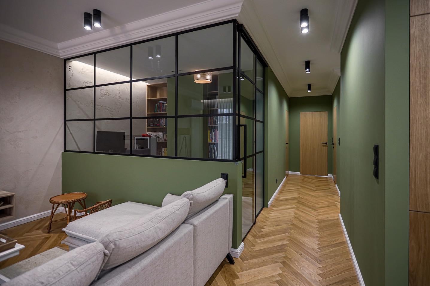 Nowoczesne wnętrze z podziałem pokoju za pomocą szklanej ściany z czarnymi szprosami, parkiet ułożony w jodełkę, ściany w odcieniach zieleni i beżu, oświetlenie punktowe.