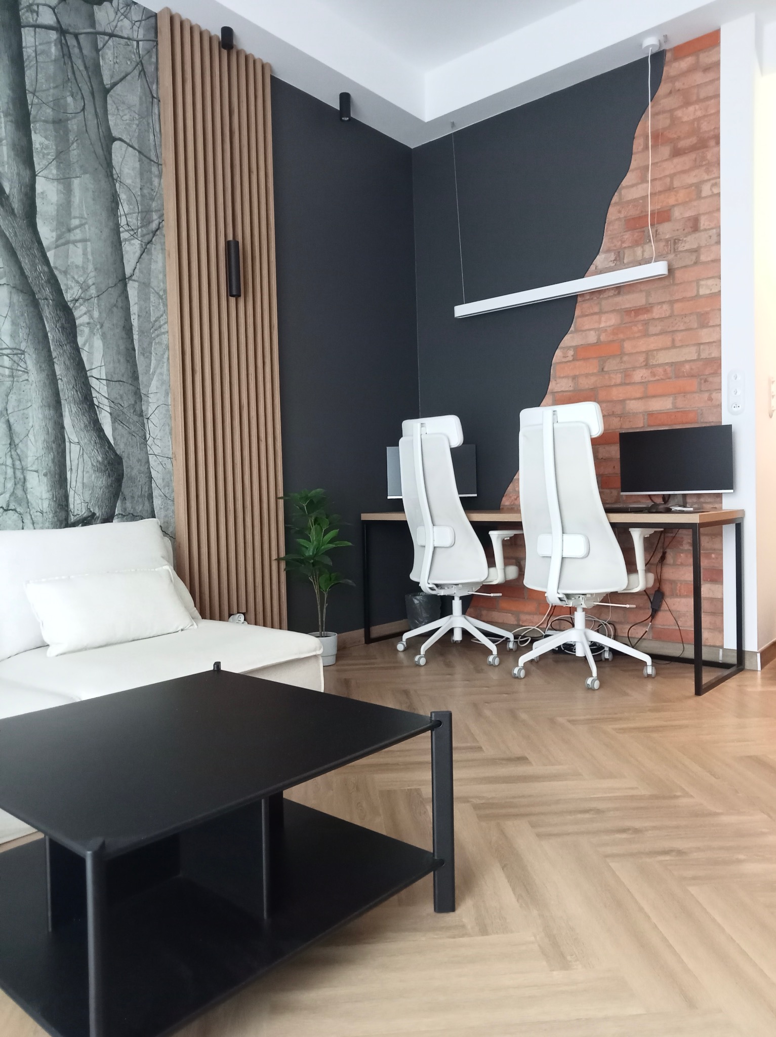 Nowoczesne wnętrze z parkietem w jodełkę, ścianą z cegły i drewnianymi lamelami, biurko z białymi krzesłami, czarny stolik kawowy i sofa z białymi poduszkami.