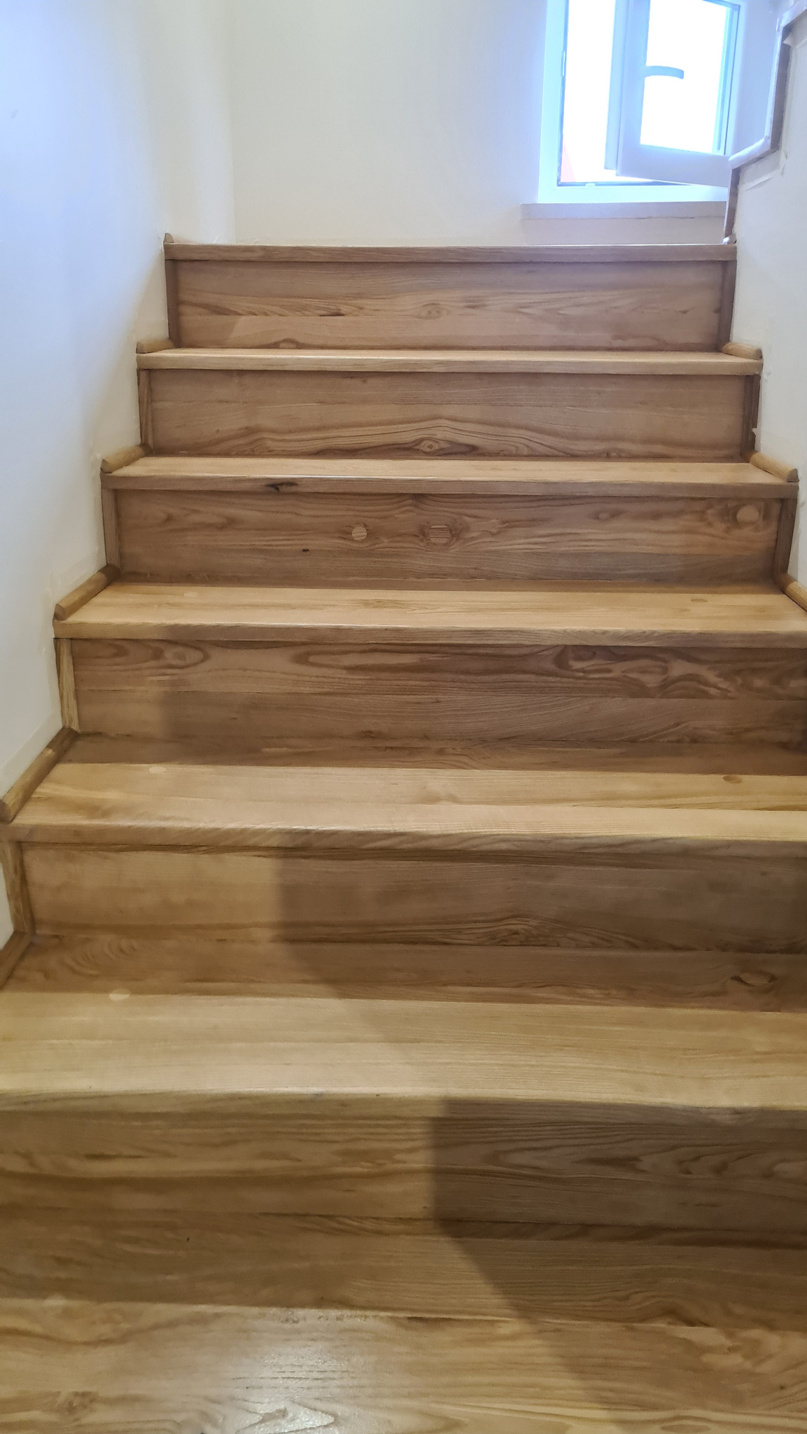 Schody drewniane z jasnego drewna, widok z dołu, świeżo po cyklinowaniu, z widocznym oknem w tle.