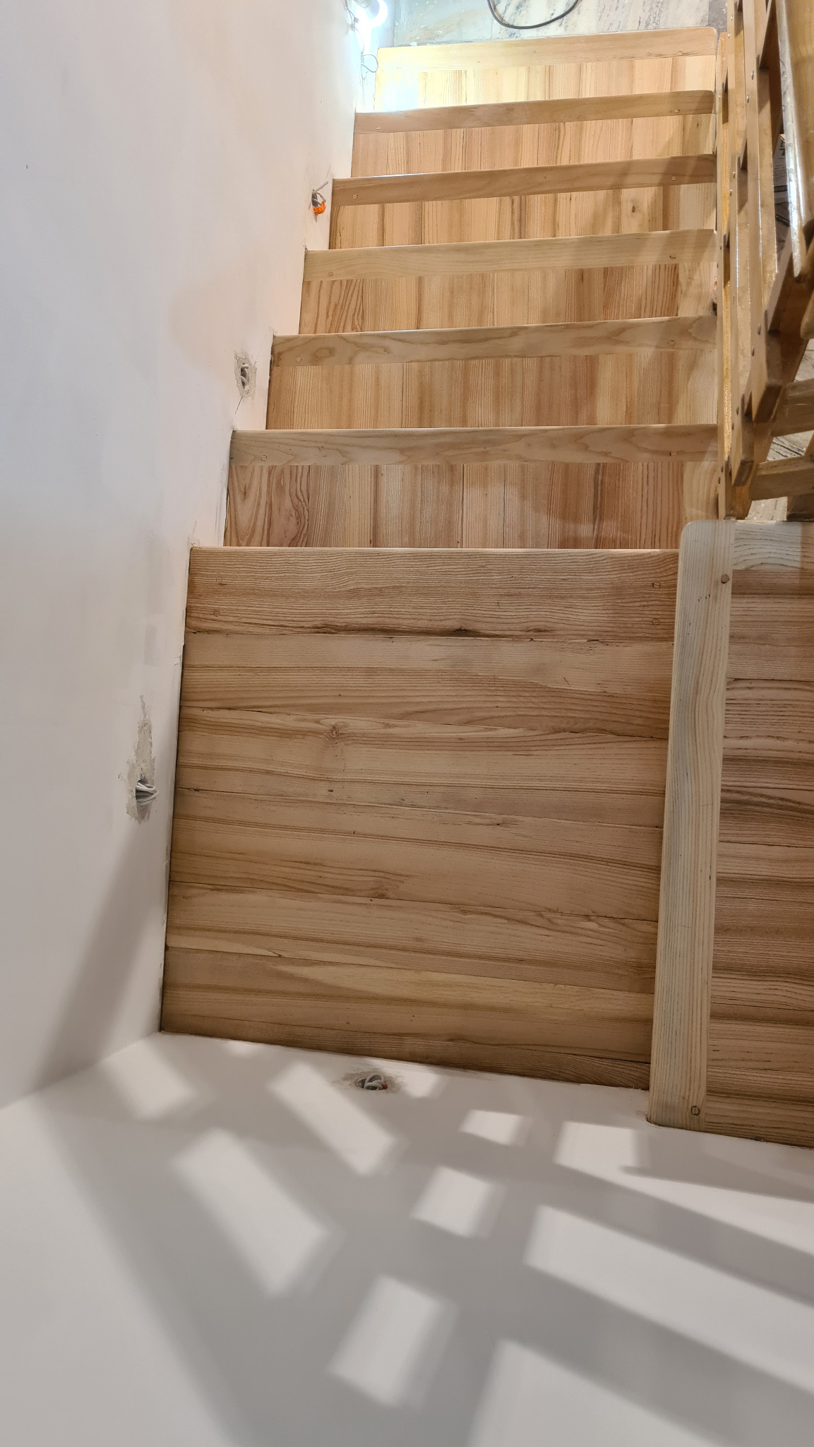 Jasne, drewniane schody w trakcie montażu, widoczne niedoskonałości na ścianie i brak wykończenia, surowe drewno balustrady.