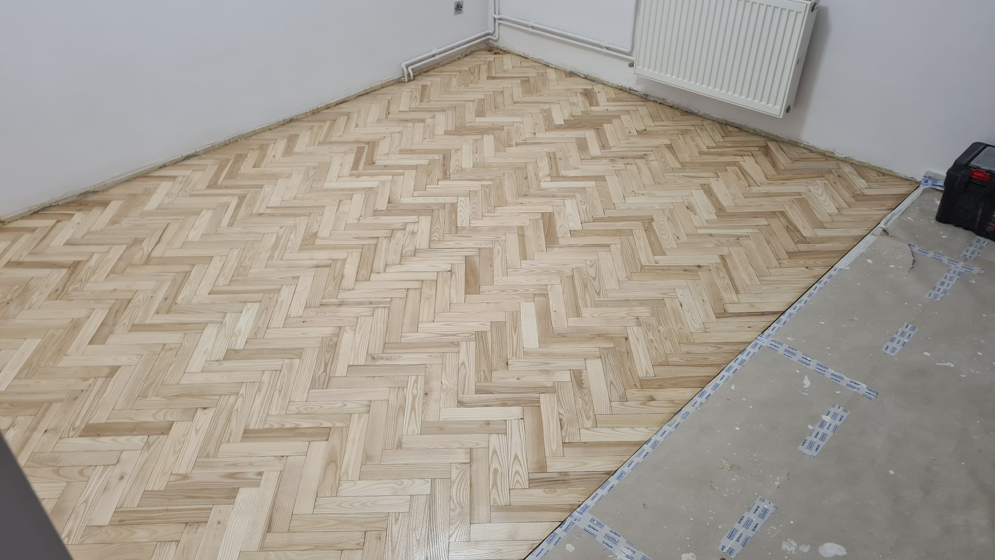 Jodełka klasyczna z jasnego drewna na podłodze w trakcie układania, widoczny fragment surowej posadzki zabezpieczonej taśmą i narzędzia w rogu pomieszczenia.