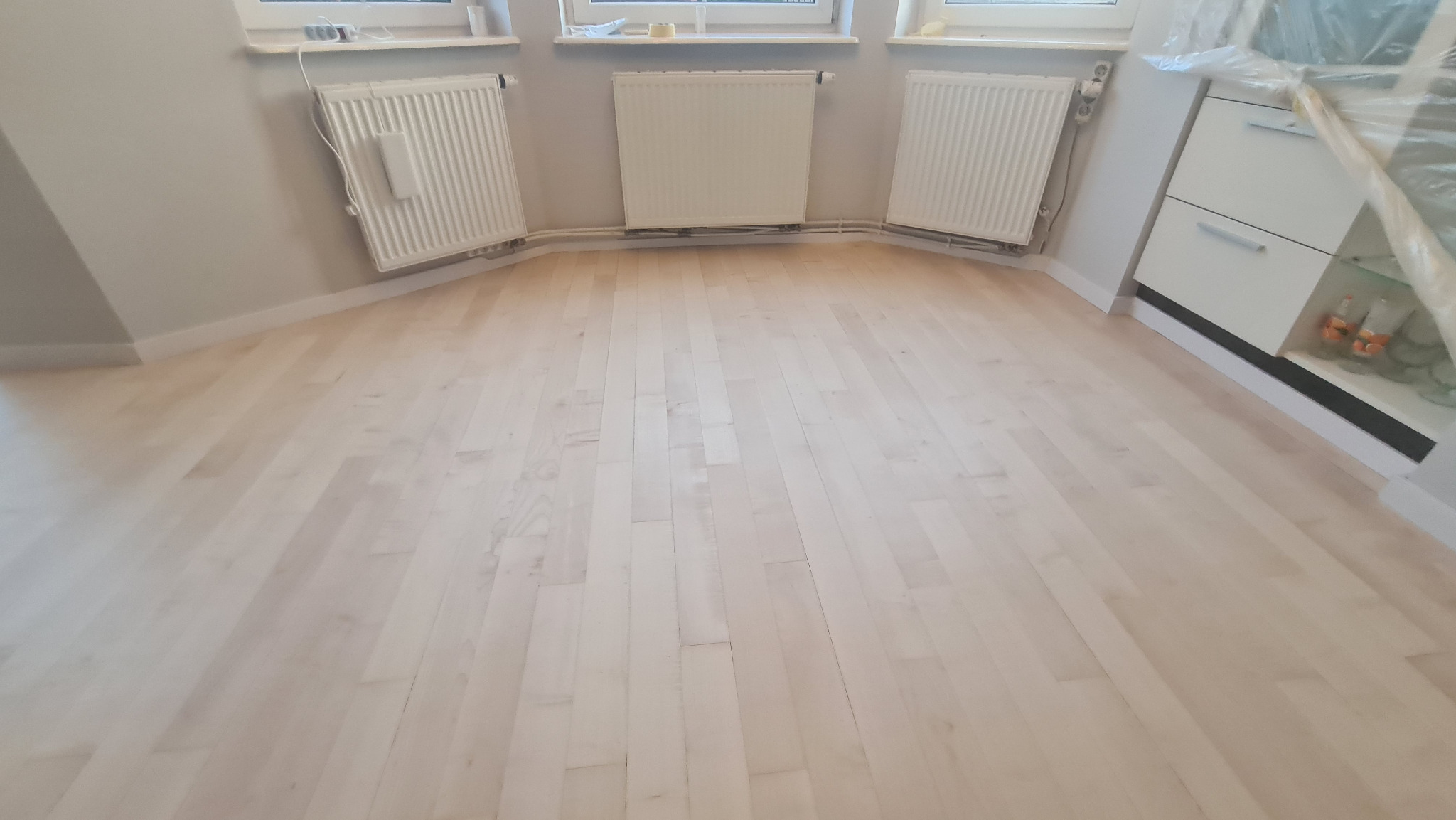 Jasna, odnowiona podłoga z parkietu w pokoju z trzema grzejnikami, widoczne ślady po cyklinowaniu na niektórych deskach, białe listwy przypodłogowe.