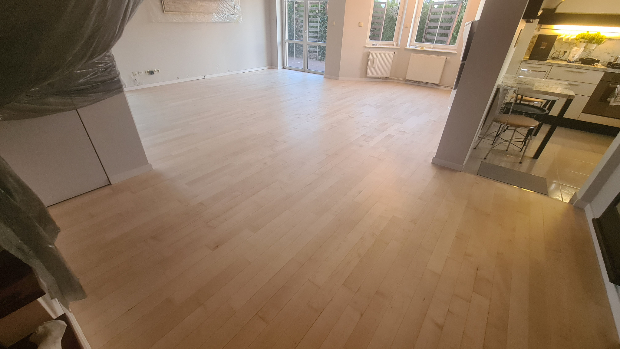 Jasne, nowo położone panele podłogowe w przestronnym salonie z widokiem na kuchnię i ogród przez duże okna balkonowe.