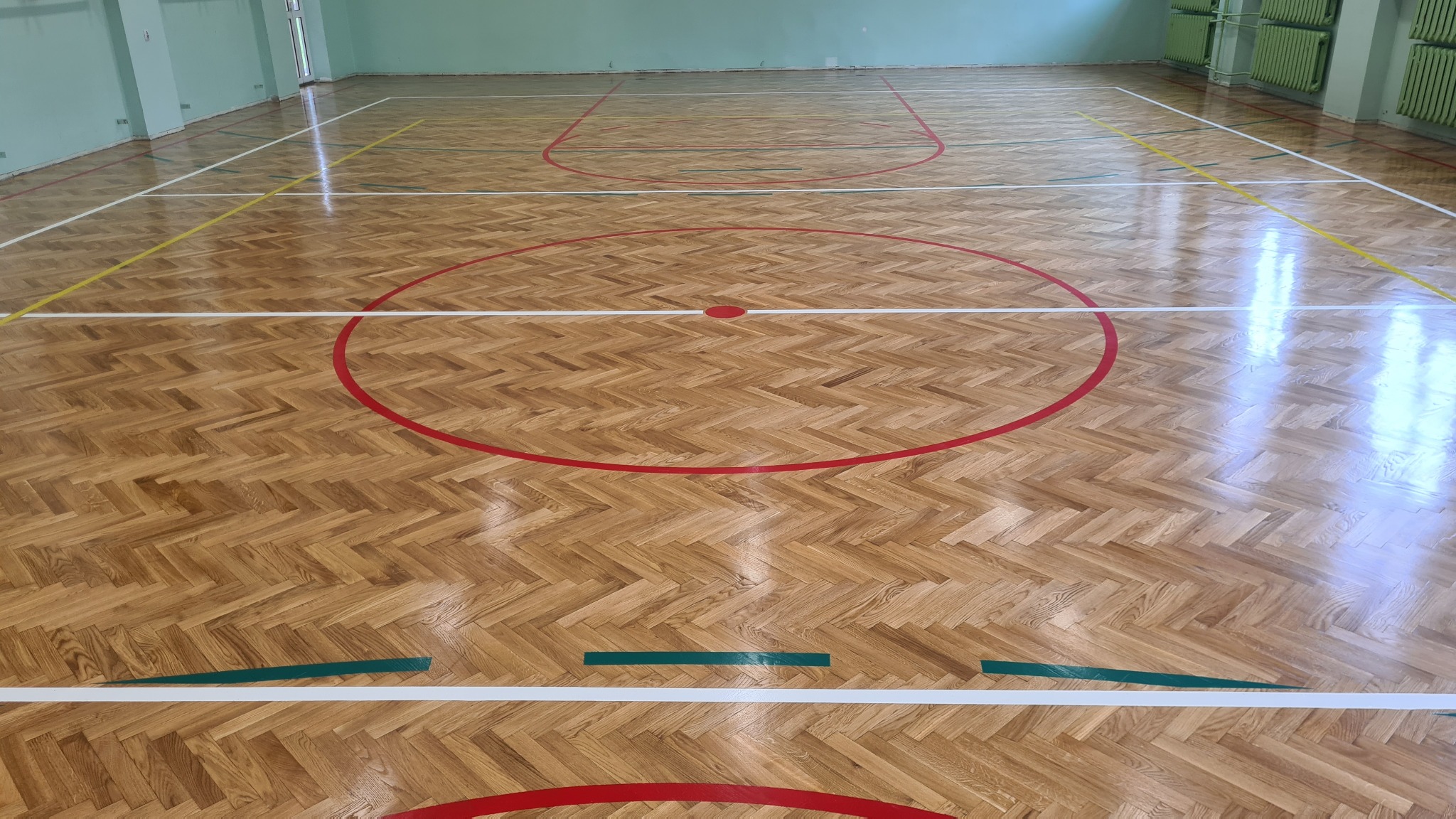 Parkiet w jodełkę na sali gimnastycznej po renowacji, z wyraźnie odświeżonymi liniami boiska w kolorach czerwonym, białym, żółtym i zielonym. Widok z góry.