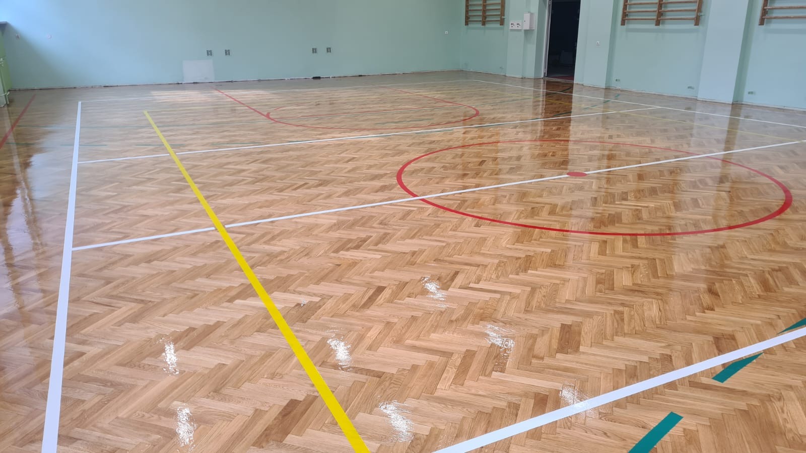 Błyszcząca, odnowiona parkietowa podłoga sali gimnastycznej z wyraźnie odmalowanymi liniami boiska w kolorach: białym, żółtym, czerwonym i zielonym.