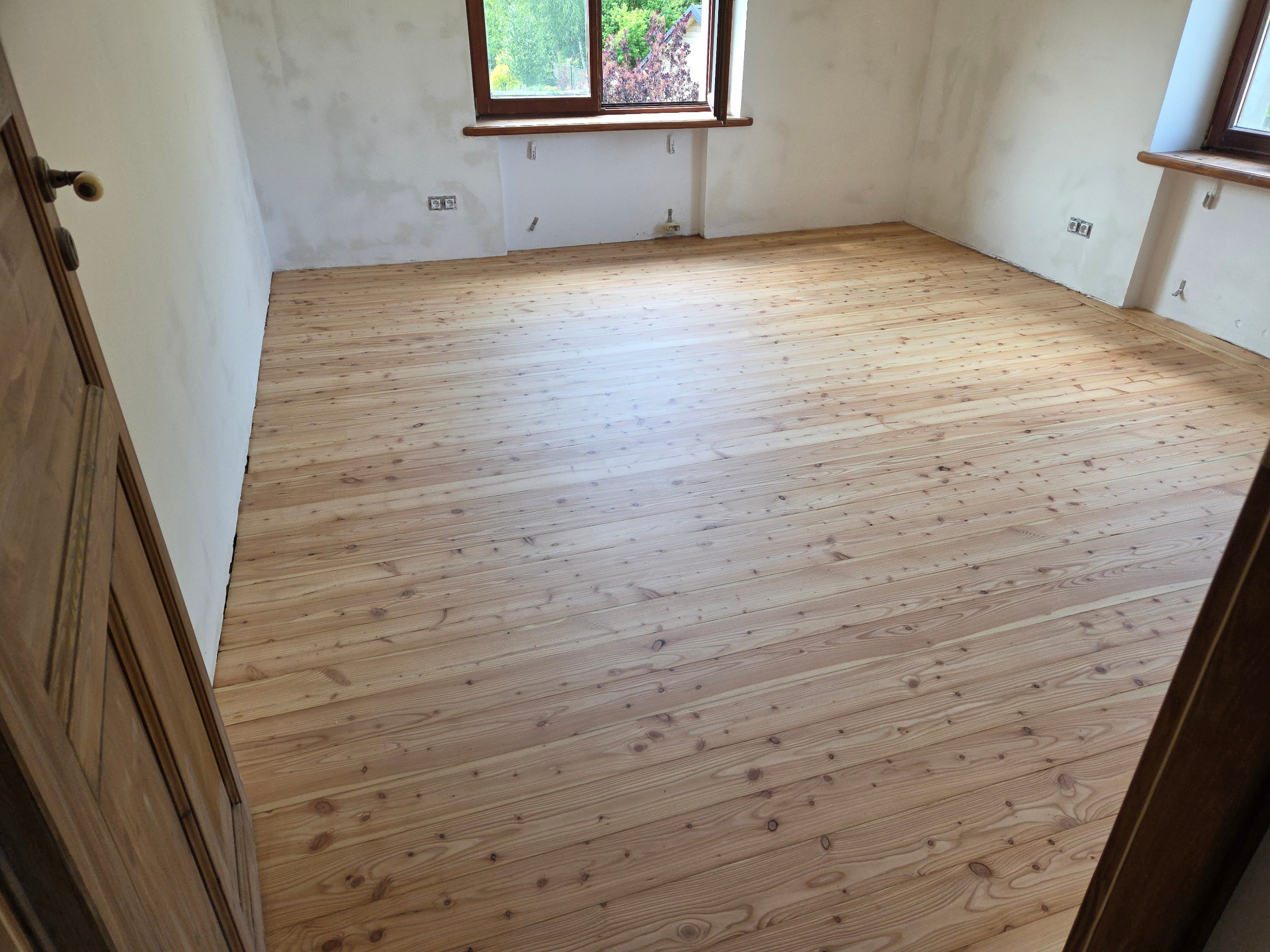 Jasna, odnowiona drewniana podłoga w pokoju z oknem. Widoczne ślady po szpachlowaniu na ścianach i drewniane drzwi po lewej stronie. Naturalne oświetlenie.