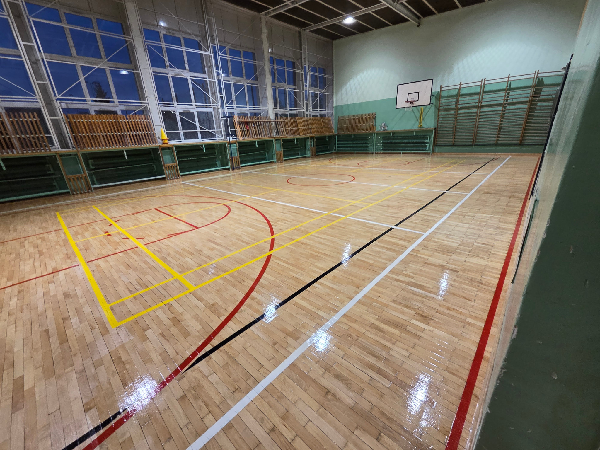 Sala gimnastyczna z parkietem po renowacji. Widoczne linie boiska w kolorach: żółtym, czerwonym, białym i czarnym. Parkiet błyszczący, ściany zielone.