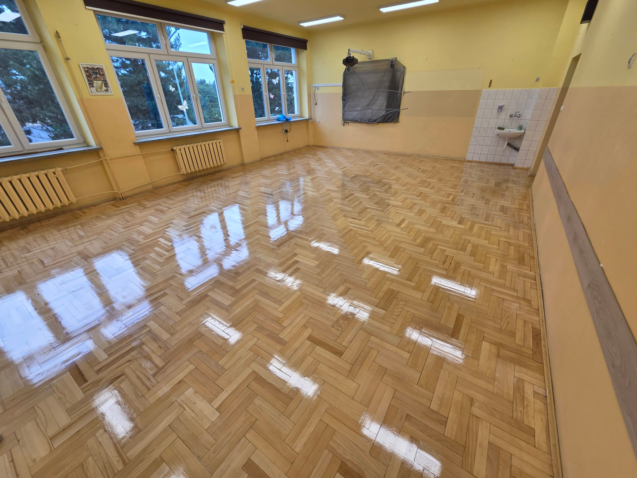 Błyszcząca, odnowiona podłoga parkietowa w jodełkę w jasnym pomieszczeniu z oknami i grzejnikami. Widoczny fragment umywalki w rogu. Świeżo położony lakier.