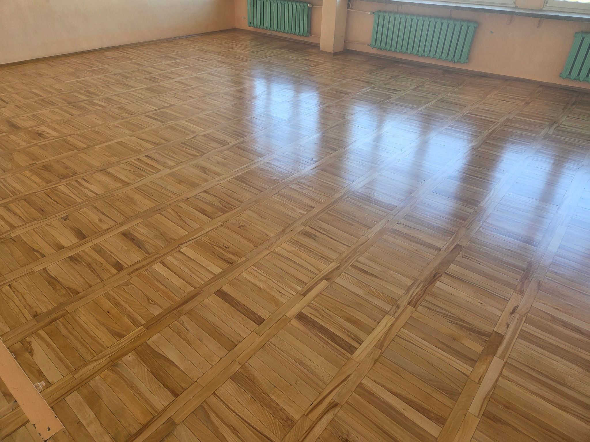 Parkiet w jodełkę po renowacji, z widocznymi refleksami światła, na tle ściany z grzejnikami w kolorze miętowym. Układ parkietu tworzy wzór prostokątów.