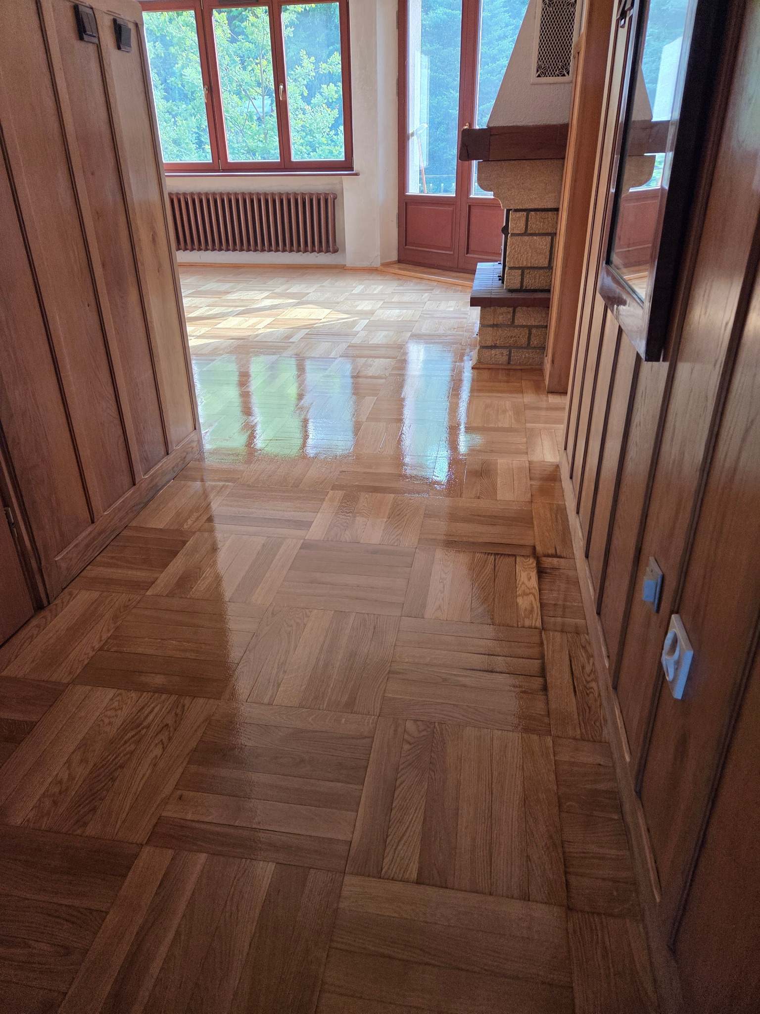 Parkiet ułożony w kwadraty, po renowacji, z widocznym połyskiem. W tle okno i grzejnik. Ściany wykończone drewnianą boazerią.