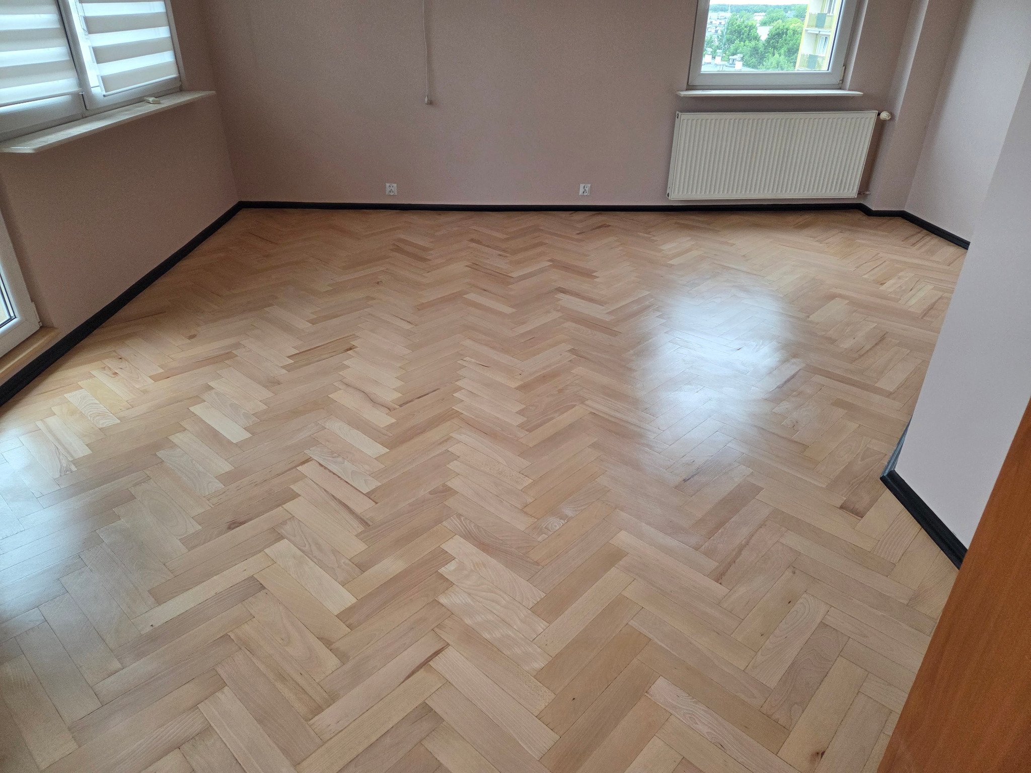 Jasna, odnowiona podłoga parkietowa w jodełkę francuską, widok na pokój z oknem i grzejnikiem. Listwy przypodłogowe w kolorze czarnym.