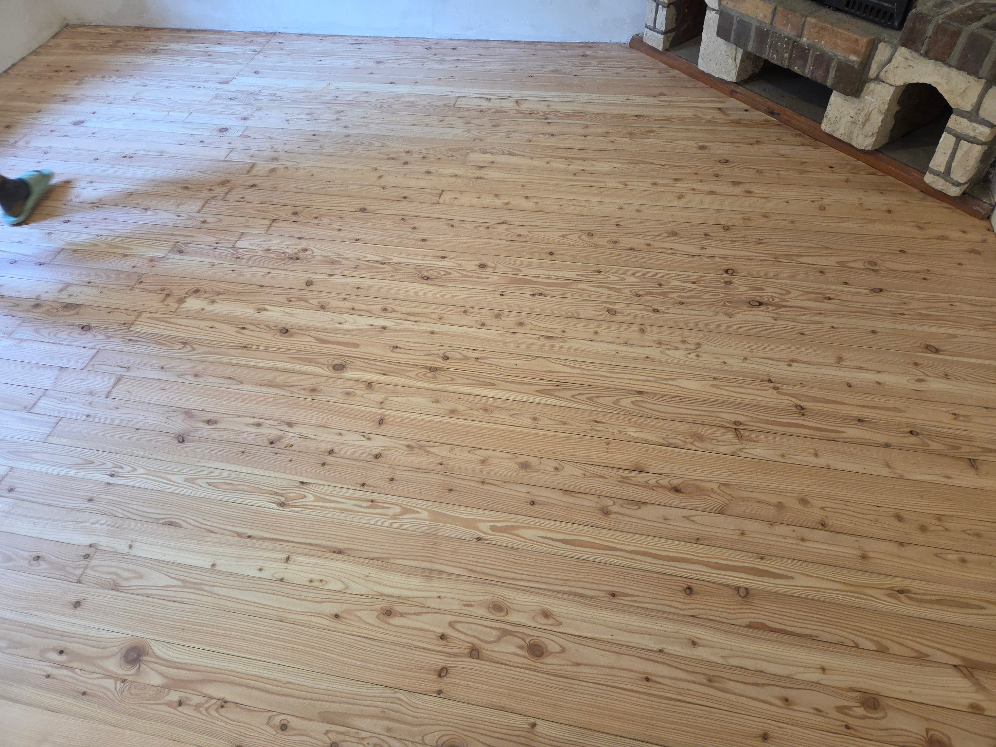 Jasna, odnowiona podłoga drewniana z widocznym wzorem słojów, fragment kominka z cegły w rogu. Świeżo wycyklinowana podłoga po renowacji, gotowa do użytku.