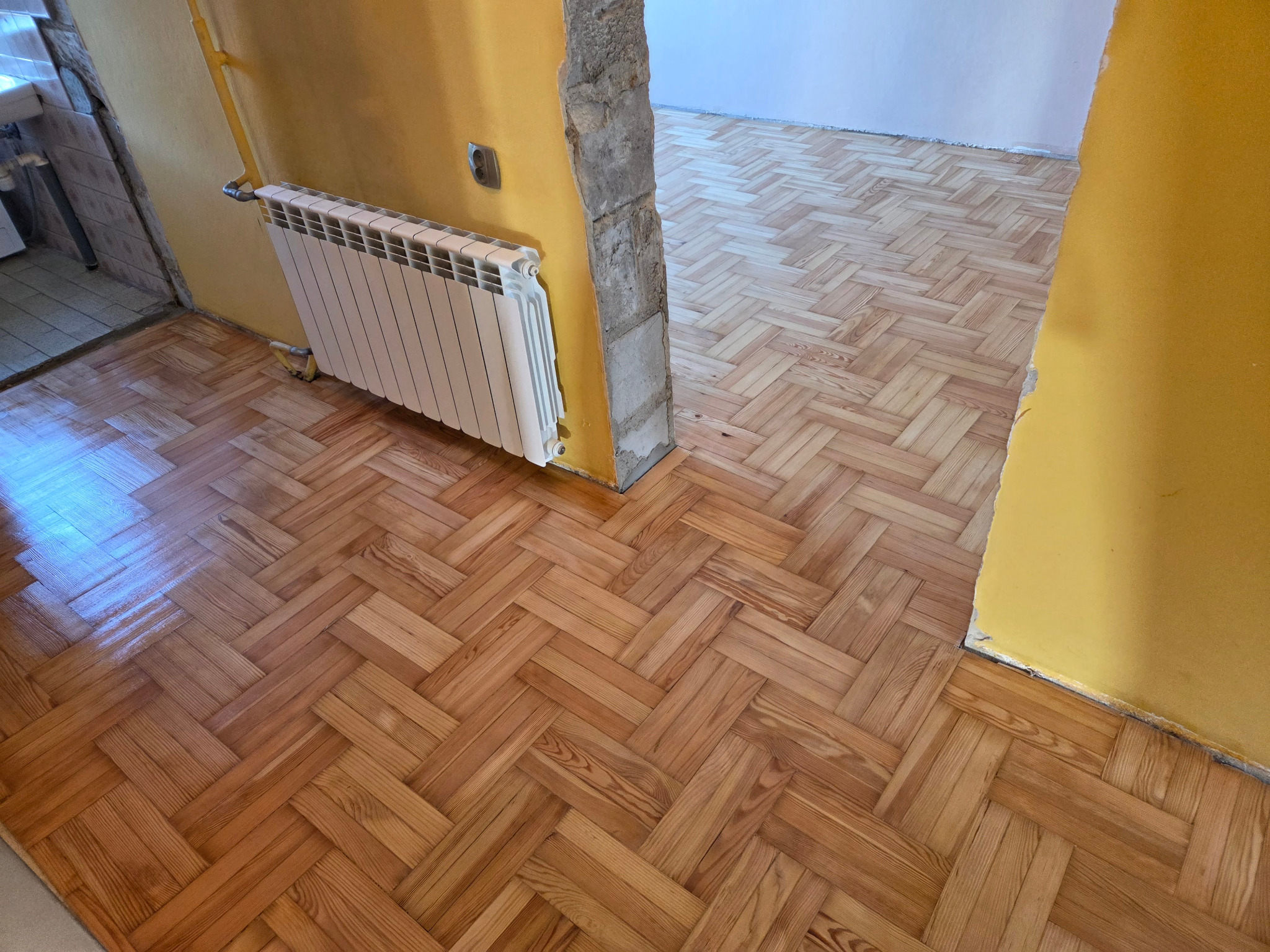 Parkiet ułożony w jodełkę, po renowacji. Widoczny grzejnik i fragment żółtej ściany. Podłoga po zabiegach odzyskuje blask i naturalny kolor drewna.
