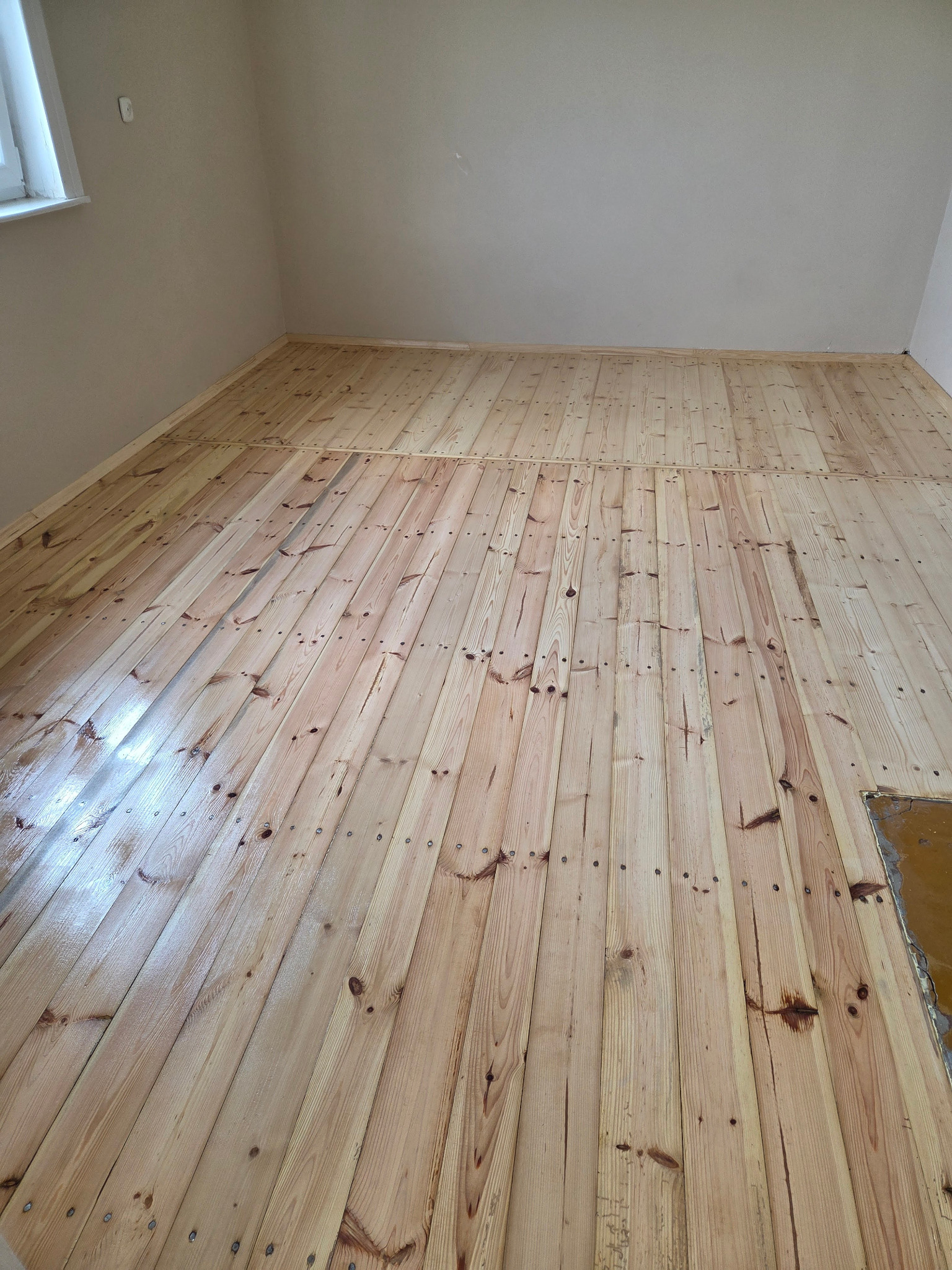 Jasna, odświeżona drewniana podłoga po renowacji, widoczne ślady po gwoździach. Fragment starej podłogi w prawym dolnym rogu. Ściany pomalowane na beżowo.