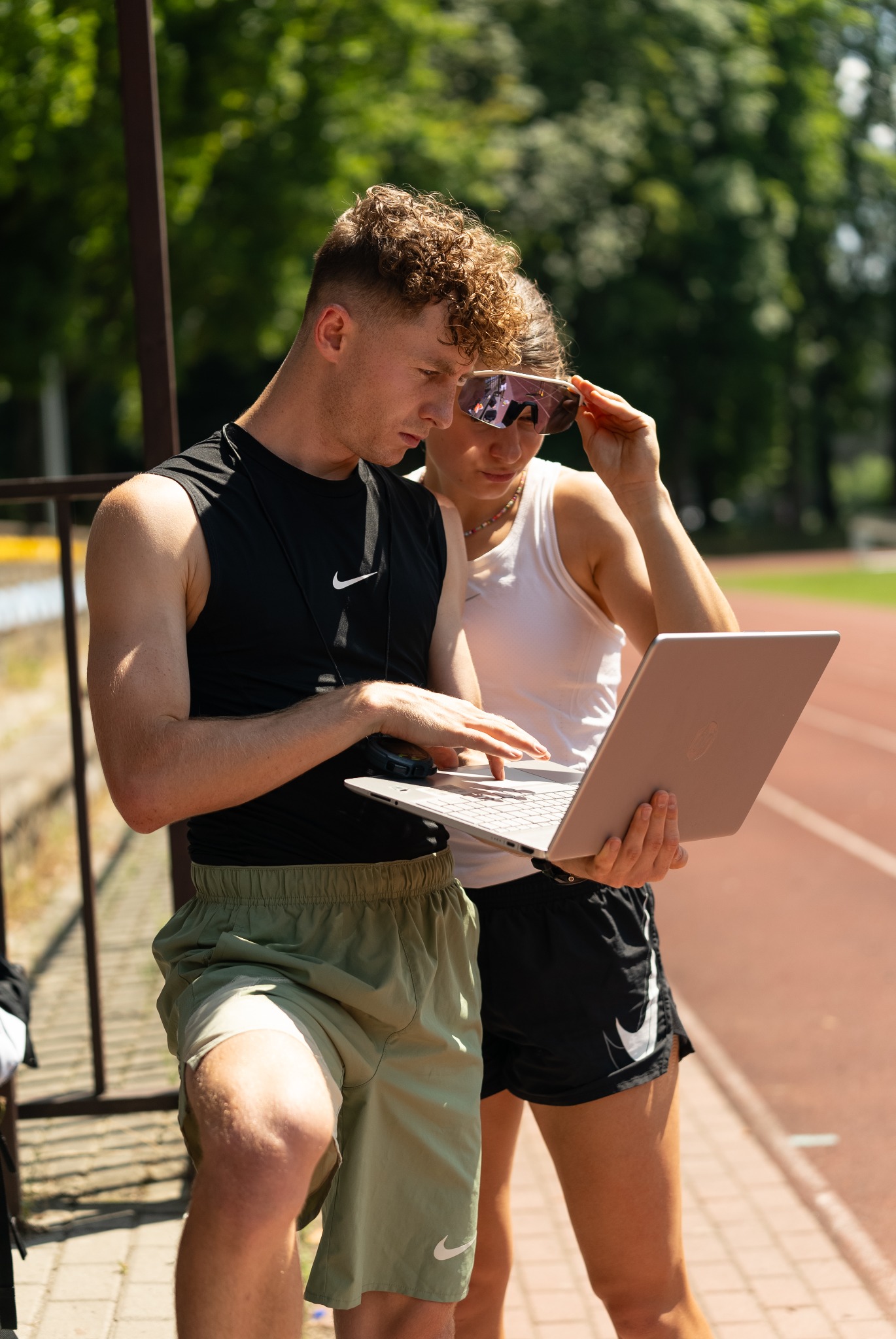 Dwoje sportowców przegląda dane treningowe na laptopie na bieżni, kobieta poprawia okulary przeciwsłoneczne.