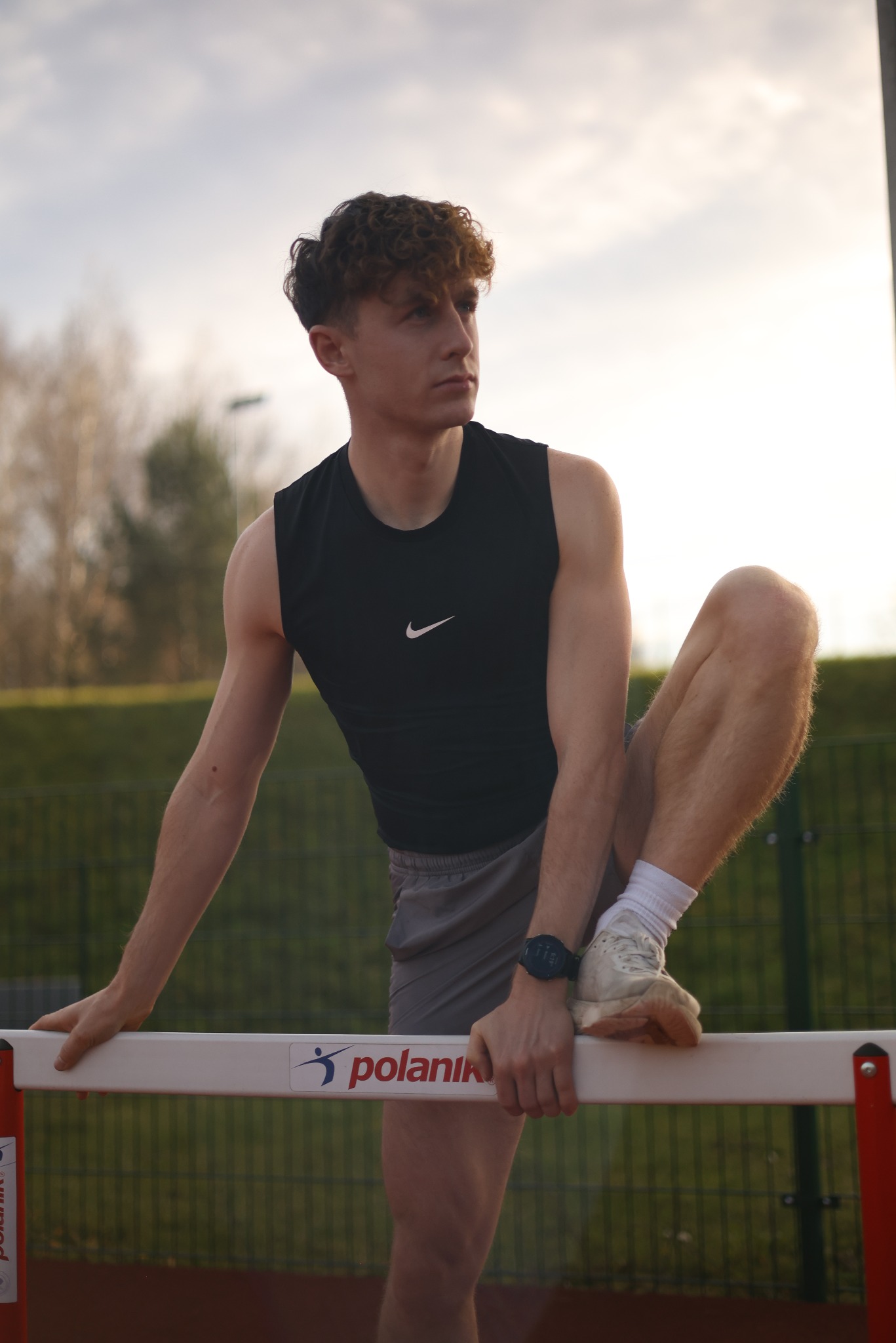 Młody mężczyzna w sportowym stroju opiera się o płotek na bieżni, z zegarkiem sportowym na nadgarstku, spoglądając w dal.