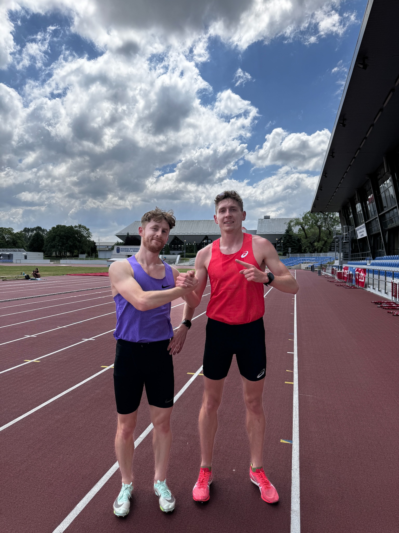 Dwóch biegaczy na bieżni stadionu, jeden w fioletowym podkoszulku i drugi w czerwonym, podają sobie ręce po treningu, w tle trybuny i pochmurne niebo.